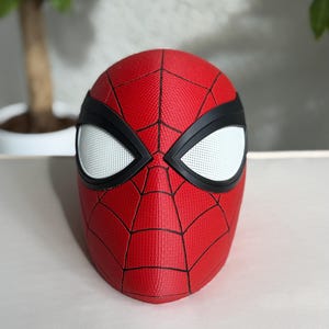 May include: A red Spider-Man mask with a textured surface and black web lines. The mask features white eye lenses outlined in black. The mask is a replica of the superhero's mask.
