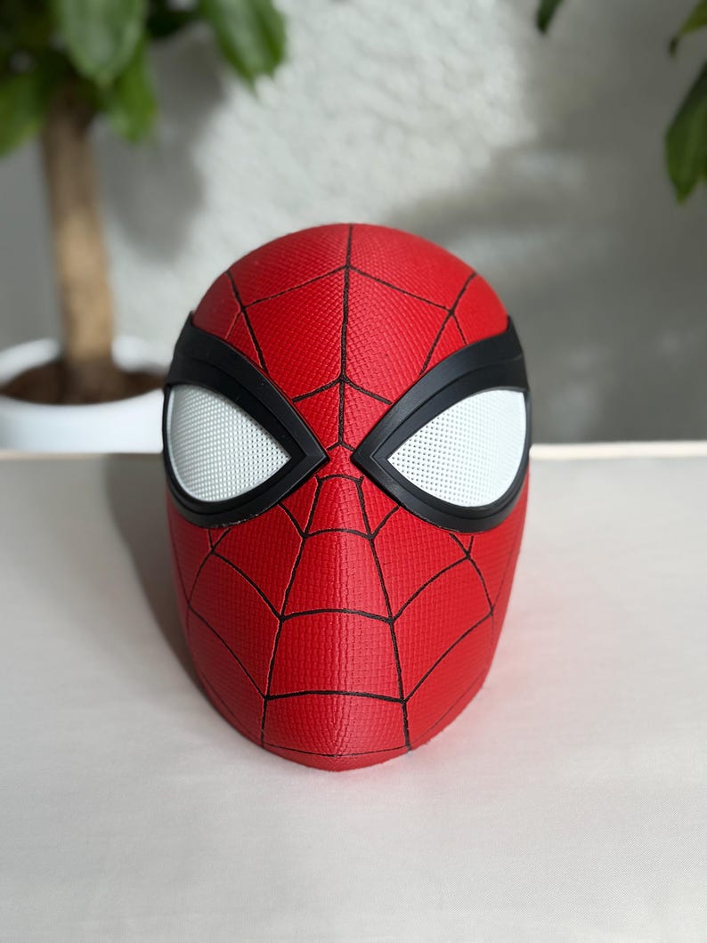 May include: A red Spider-Man mask with a textured surface and black web lines. The mask features white eye lenses outlined in black. The mask is a replica of the superhero's mask.