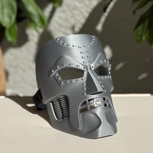 May include: A silver-colored mask with a detailed, angular design. The mask features eye openings, a prominent nose piece, and a mouth area with a mechanical appearance. It has a strap for securing it to the head, and is likely a costume or cosplay item.