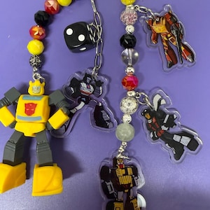 May include: A collection of keychains featuring cartoon robot characters, dice, and beaded accents. The keychains include a yellow and gray robot figure, and several clear acrylic charms with robot designs. The keychains are attached to a black ring.