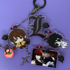 May include: A keychain featuring anime-style character charms. The charms include a large black letter "L", a character with brown hair, and a character with red hair. The keychain also has a small apple charm and a cross charm.