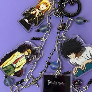 May include: Keychain featuring anime character charms, a black pom-pom, and decorative chains. The charms depict characters from Death Note. The keychain includes a small black book charm with the words "Death Note".