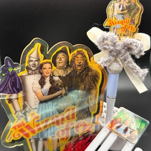 May include: A Wizard of Oz themed acrylic display featuring Dorothy, the Tin Man, Scarecrow, and Cowardly Lion, with a separate pen topped by a small character cutout and a white U-shaped paperclip accessory with a square image of the cast.