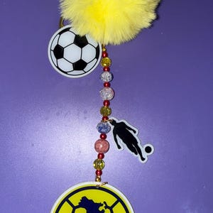 Puede incluir: Un llavero con temática de fútbol que presenta un pompón amarillo, un balón de fútbol, una silueta de futbolista y un emblema redondo con las letras "CA" y un mapa de América. El llavero tiene una cadena de cuentas.