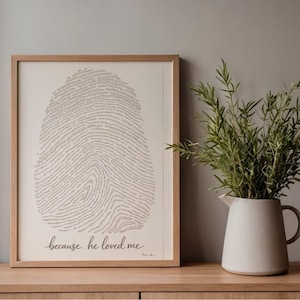 May include: A framed print with a fingerprint design made of text, in a light wood frame. The print has the cursive phrase "because he loved me". A white jug with greenery is to the right, on a wooden surface.