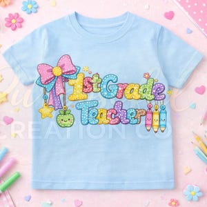 Può includere: Una maglietta azzurra con la scritta "1st Grade Teacher" in lettere colorate cucite. Il disegno comprende un fiocco, una mela, stelle e matite. La maglietta è circondata da decorazioni a tema scolastico.