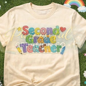 Può includere: T-shirt color crema con la scritta "Second Grade Teacher" in lettere colorate cucite. Il design include una stella, un cuore, una matita e un pastello. La maglietta è esposta su una superficie verde con decorazioni a tema scolastico.