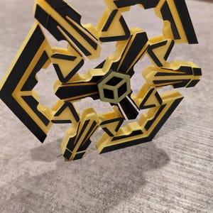 May include: A geometric, three-dimensional object with a complex, open design. The object features a repeating pattern of yellow and black angular shapes, with a central cube-like structure. The overall form is symmetrical and appears to be made of plastic.
