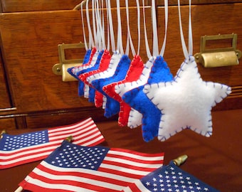 May include: Handmade felt star ornaments in red, white, and blue, hanging from white ribbons. The stars are stitched with contrasting thread. Small American flags are displayed below, creating a patriotic theme. These ornaments are perfect for Independence Day.