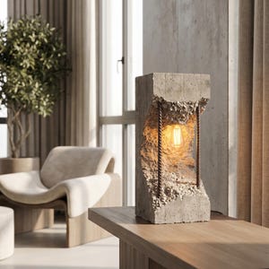 May include: A modern concrete table lamp with a rough, textured surface and a warm, glowing lightbulb. The lamp features a unique design with exposed metal wiring and sits on a wooden table, showcasing an industrial aesthetic.