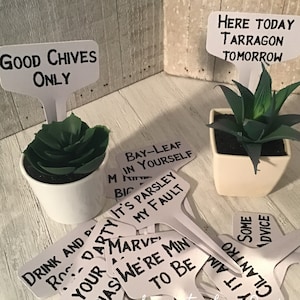 May include: A collection of white plant markers with black handwritten text. The text includes phrases like "Good Chives Only", "Here Today Tarragon Tomorrow", "Bay-Leaf Min Yourself", "It's Parsley My Fault", "Drink and Party", "We're Min To Be", "Say It Aim Some Advice", and "Cilantro". The markers are arranged on a white wooden surface with two small potted succulents.
