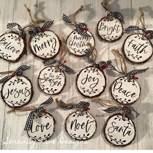 May include: Twelve round wood slices with white paint and black lettering. Each slice has a different Christmas-themed word or phrase, such as "Merry", "Peace", "Joy", "Believe", "Faith", "Santa", "Noet", "Love", "Jesus", "Tis the Season", "Bright", and "Merry Christmas".