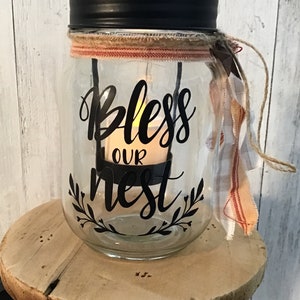 May include: A glass jar with a black lid and a burlap ribbon around the top. The jar has a black vinyl decal that says "Bless Our Nest" with a small candle inside.