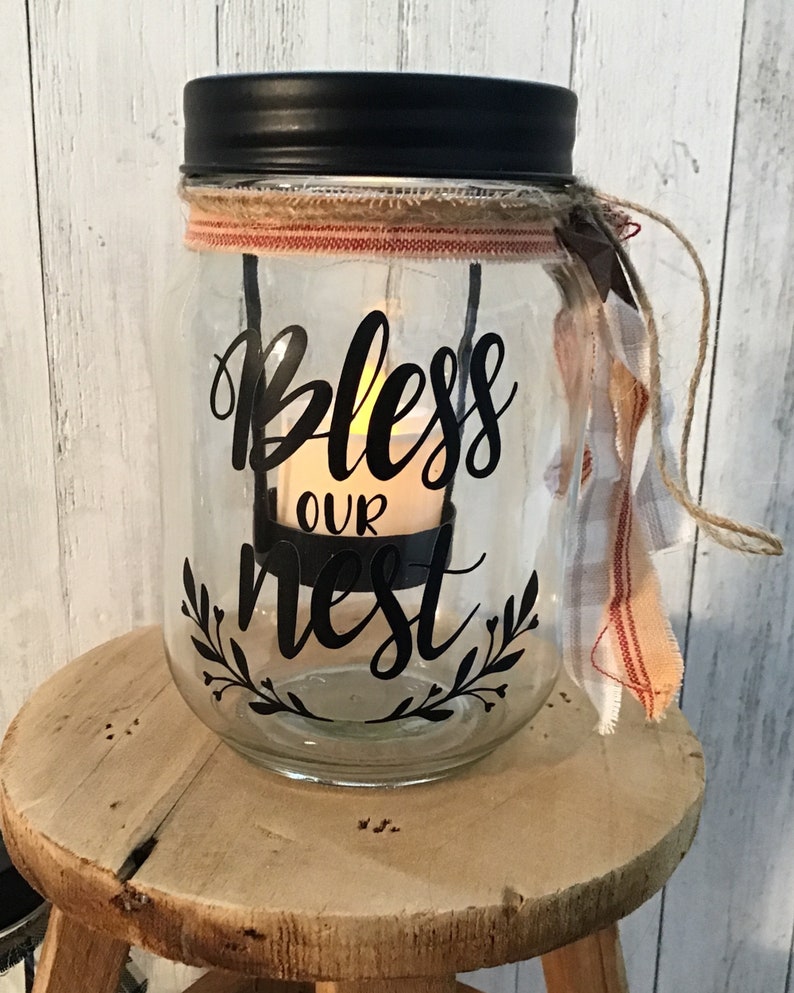 May include: A glass jar with a black lid and a burlap ribbon around the top. The jar has a black vinyl decal that says "Bless Our Nest" with a small candle inside.