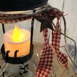 May include: A glass jar candle lantern with a black lid, red and white gingham ribbon, and a rustic metal star. The jar has a black metal ring around the top and a black metal base. The candle is lit and the jar is decorated with the words "Bless this home".