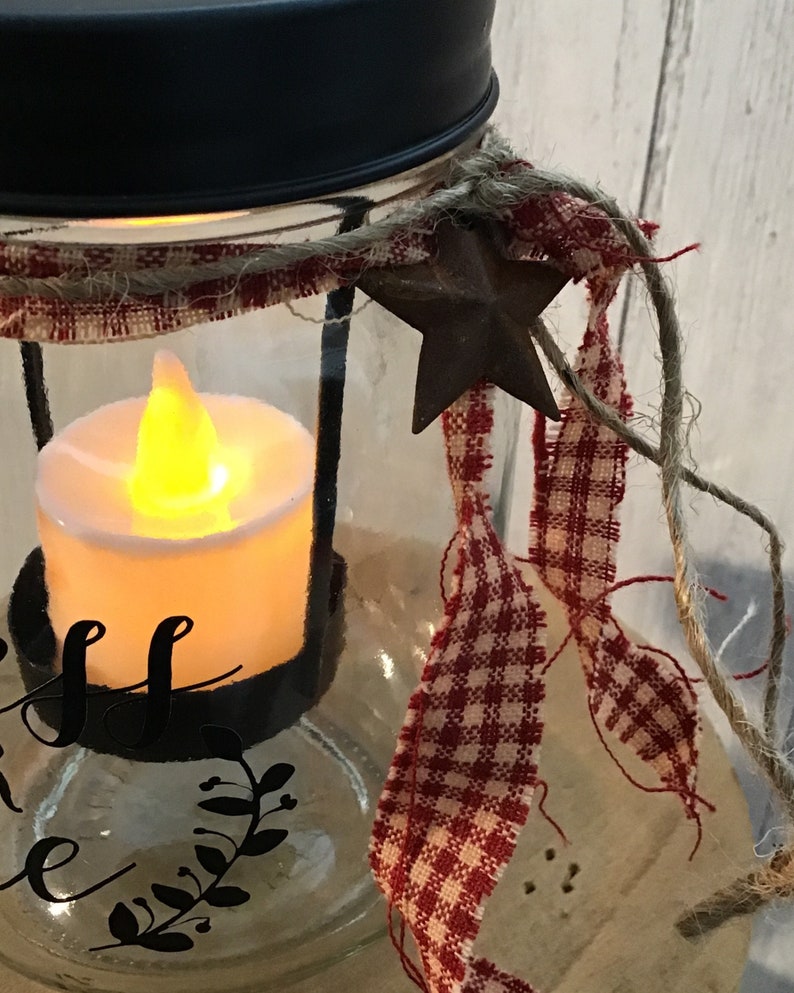 May include: A glass jar candle lantern with a black lid, red and white gingham ribbon, and a rustic metal star. The jar has a black metal ring around the top and a black metal base. The candle is lit and the jar is decorated with the words "Bless this home".