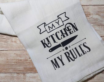May include: White kitchen towel with the text "MY KITCHEN MY RULES" in black. The design includes a knife graphic and banner elements. The towel is folded and lies on a light wood surface.