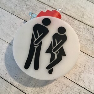 May include: A white round light switch cover with black silhouettes of two figures in a restroom pose. The switch cover has a red electrical cord and plug.