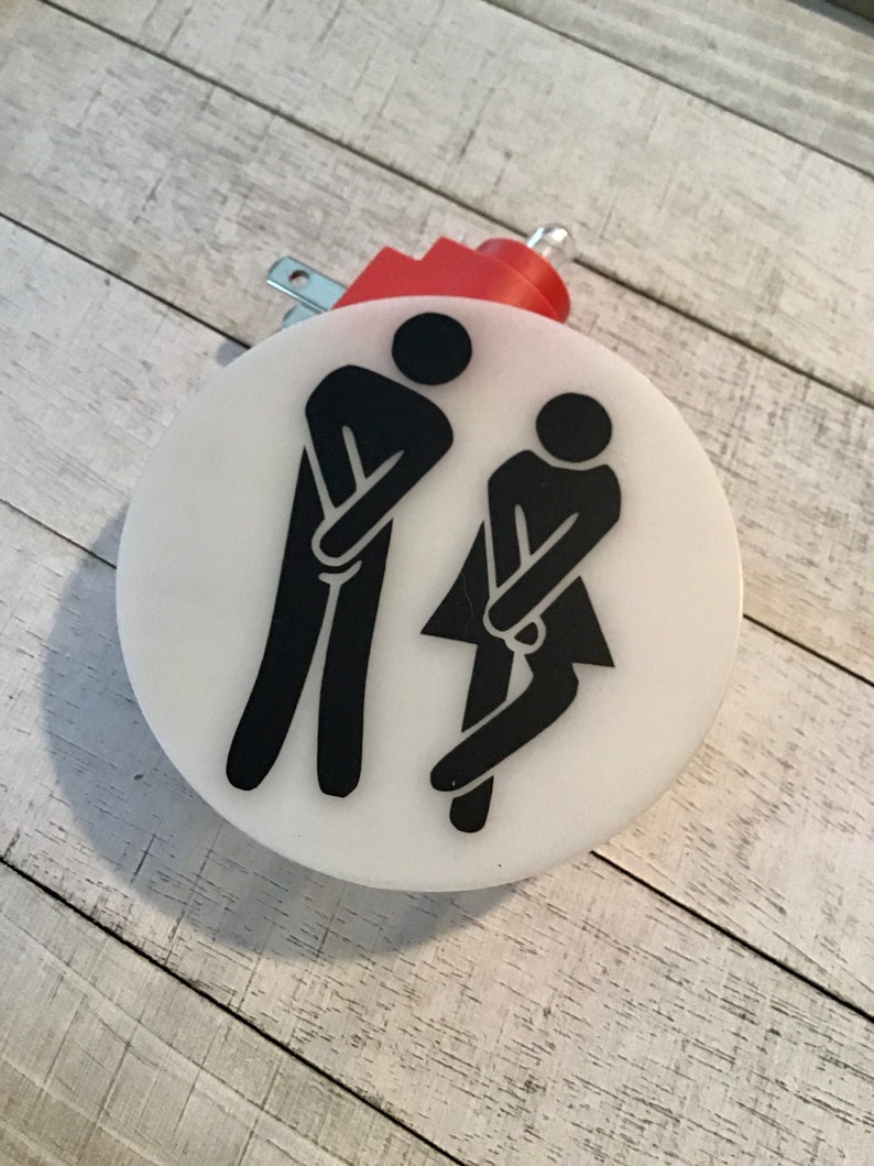 May include: A white round light switch cover with black silhouettes of two figures in a restroom pose. The switch cover has a red electrical cord and plug.