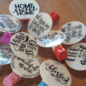 May include: A collection of 10 round white night lights with various inspirational quotes printed in black. The night lights are plugged into electrical outlets with different colored plugs.
