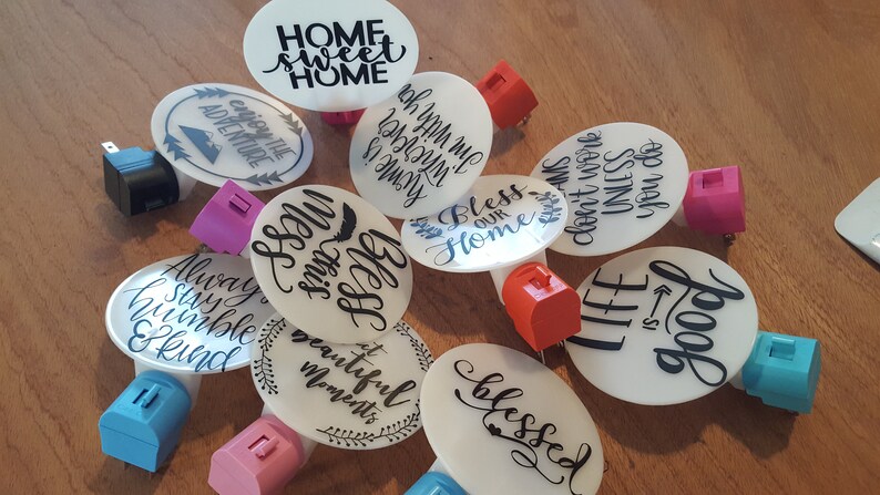 May include: A collection of 10 round white night lights with various inspirational quotes printed in black. The night lights are plugged into electrical outlets with different colored plugs.