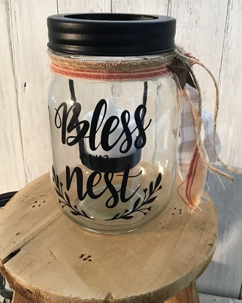 May include: A clear glass jar with a black lid and a burlap and red ribbon around the top. The jar has a black vinyl decal that says "Bless our nest" with a leafy design around the text. The jar is sitting on a wooden surface.