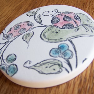 May include: A white ceramic coaster with a black and white illustration of ladybugs and leaves. The ladybugs have pink and black spots.