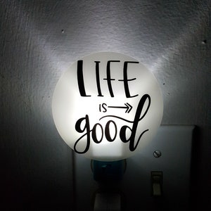 May include: White night light with the text "LIFE IS good" in black lettering. The light is plugged into an electrical outlet.
