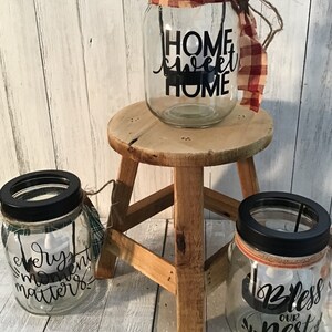 May include: Three glass jars with black lids and decorative sayings. The jars are decorated with plaid fabric and twine. The sayings on the jars are "Home Sweet Home", "Every Moment Matters", and "Bless Our Nest".