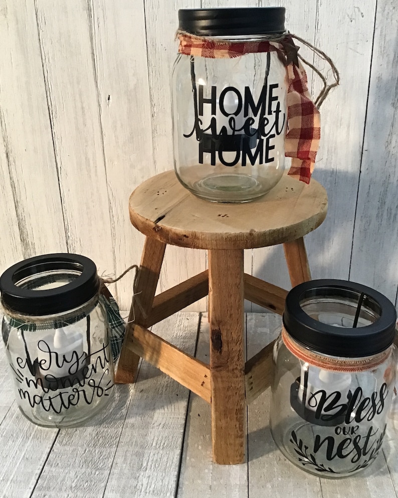 May include: Three glass jars with black lids and decorative sayings. The jars are decorated with plaid fabric and twine. The sayings on the jars are "Home Sweet Home", "Every Moment Matters", and "Bless Our Nest".