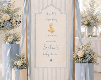 May include: A vertical rectangular banner featuring a light beige background with thin blue vertical stripes. The text reads A Little Duckling IS ON THE WAY welcome to Sophia's baby shower, accompanied by a small yellow duckling and a blue heart.