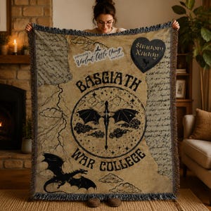 Puede incluir: Una manta beige con dise&ntilde;os de dragones y texto en negro. La manta presenta las palabras "Basgiath War College" y "Shadow Daddy". Tiene un borde con flecos y una est&eacute;tica vintage.
