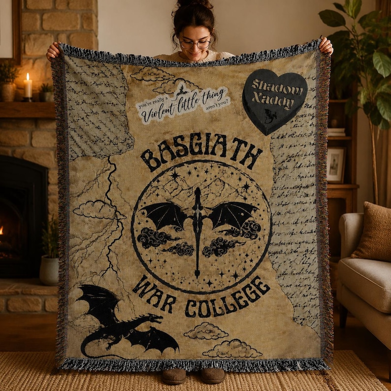 Puede incluir: Una manta beige con dise&ntilde;os de dragones y texto en negro. La manta presenta las palabras "Basgiath War College" y "Shadow Daddy". Tiene un borde con flecos y una est&eacute;tica vintage.