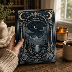 Op de afbeelding: Een donkerblauwe e-readerhoes met een goudkleurige illustratie van bergen, een meer en een maansikkel. De tekst luidt: "To the stars who listen, and the dreams that are answered." Het ontwerp bevat zwaarden, bloemmotieven en sterren.
