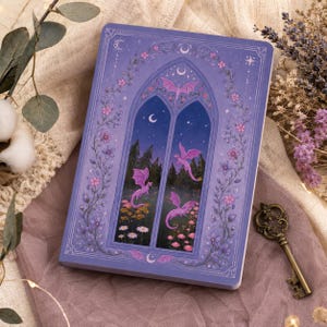 Op de afbeelding: Een paars notitieboek met een fantasie-illustratie. De omslag toont een vensterscène met roze draken, bloemen en een nachthemel. Het notitieboek is omgeven door gedroogde lavendel, een decoratieve sleutel en een neutraal gekleurde stof.