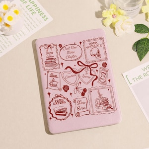 Puede incluir: Una funda rosa claro para lector de libros electrónicos con varias ilustraciones en rosa oscuro. Las ilustraciones incluyen libros, corazones, lazos y las frases "Just One More Chapter" y "Dark Romance".