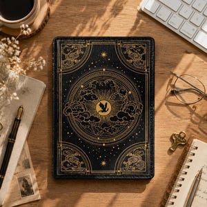 Könnte beinhalten: Schwarzes Notizbuch mit einem goldenen Himmelsdesign mit einem Drachen, Wolken und Sternen. Das Notizbuch hat eine lederartige Textur und liegt auf einem Holztisch mit Tastatur, Brille und einer Tasse Kaffee.