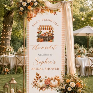 May include: A vertical rectangular banner with a light orange gingham border, featuring the text "SHE'S FRESH OFF the market WELCOME TO Sophia's BRIDAL SHOWER". The design includes a market stall illustration, pumpkins, and autumn flowers.
