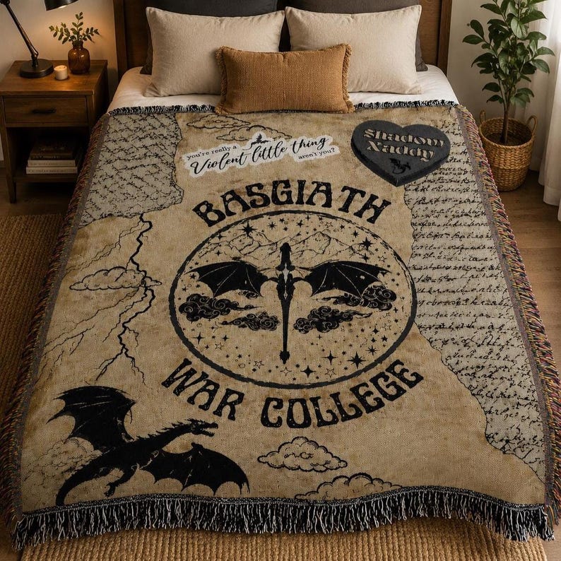 Puede incluir: Manta tejida beige con un gr&aacute;fico de drag&oacute;n negro y las palabras "BASGIATH WAR COLLEGE". La manta presenta un ribete de flecos y texto adicional, incluyendo "Violent little thing aren't you?" y "Shadow Daddy".