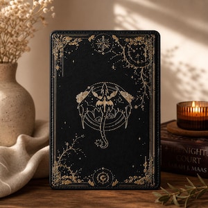 Puede incluir: Cubierta de libro negra con detalles dorados. El diseño presenta una ilustración central de una criatura alada, rodeada de motivos celestiales y florales. El título no es visible.