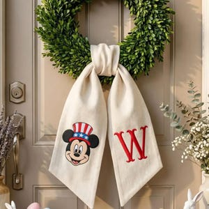 Op de afbeelding: Een groene krans hangt aan een deur, versierd met een crèmekleurige stoffen sjerp. Eén kant heeft een Mickey Mouse-ontwerp in patriottische kleuren, de andere een rode "W". De deur heeft een zilveren handvat en slot.