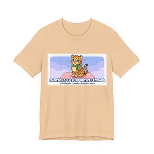 May include: A pale beige t-shirt with a cartoon leopard wearing a green scarf, sitting on a pink rug. The text "Emotionally Supportive Leopard" and "Certified in Comfort & Mild Chaos" are printed on the shirt.