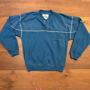 May include: A blue long-sleeve pullover shirt featuring a waffle-knit texture on the lower body and sleeves, a smooth upper chest panel, a three-button placket, a single chest pocket, and white contrast stitching throughout the garment.