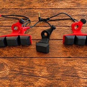 May include: Two red and black mechanical keyboard switch-style triggers with black cables and gold-plated audio jacks, accompanied by a single black trigger component, all featuring a distinct blocky, keycap-like design.