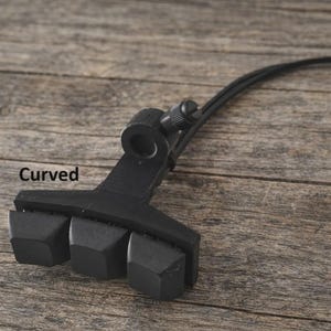 May include: A black, curved support with three angled pads, a circular opening, and a screw adjustment. The word "Curved" is printed on the surface. The support has a flexible black cord attached.