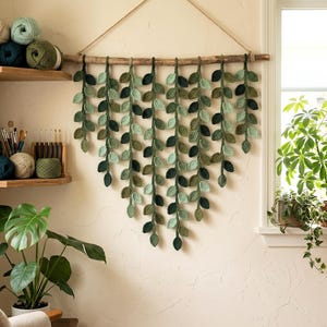 Puede incluir: Un colgante de pared hecho a mano con hojas verdes en cascada. Las hojas, en varios tonos de verde, están unidas a una vara de madera y suspendidas por un cordón de fibra natural. El colgante se exhibe sobre una pared de color crema.