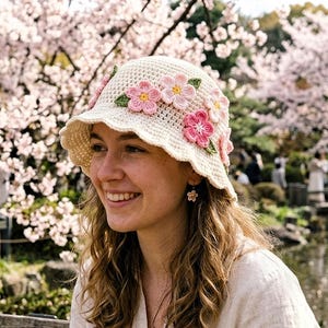 Puede incluir: Un sombrero de cubo de ganchillo color crema adornado con flores rosas y verdes. El sombrero tiene un borde festoneado y se usa al aire libre. El fondo presenta un parque con cerezos en flor.