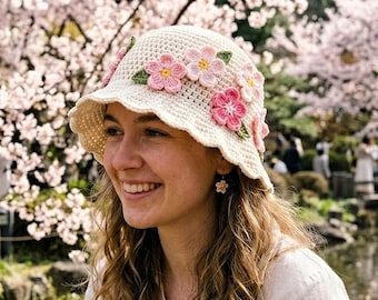 Può includere: Un cappello a secchiello all'uncinetto color crema adornato con fiori rosa e verdi. Il cappello ha un bordo smerlato e viene indossato all'aperto. Lo sfondo presenta un parco con alberi di ciliegio in fiore.