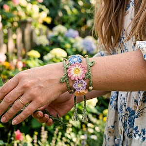 Könnte beinhalten: Ein handgefertigtes Häkelarmband mit drei bunten Blumen: eine blaue, eine rosa und eine lila Blume. Das Armband ist durch grüne Ranken mit Holzperlen und Quasten verbunden. Das Armband wird am Handgelenk getragen.