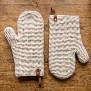 Pode incluir: Duas luvas de forno de crochê bege com laços de couro marrom. Uma é uma luva de forno completa, a outra é um pegador de panela de uma mão. As luvas são feitas de fio grosso e texturizado e são para uso na cozinha.